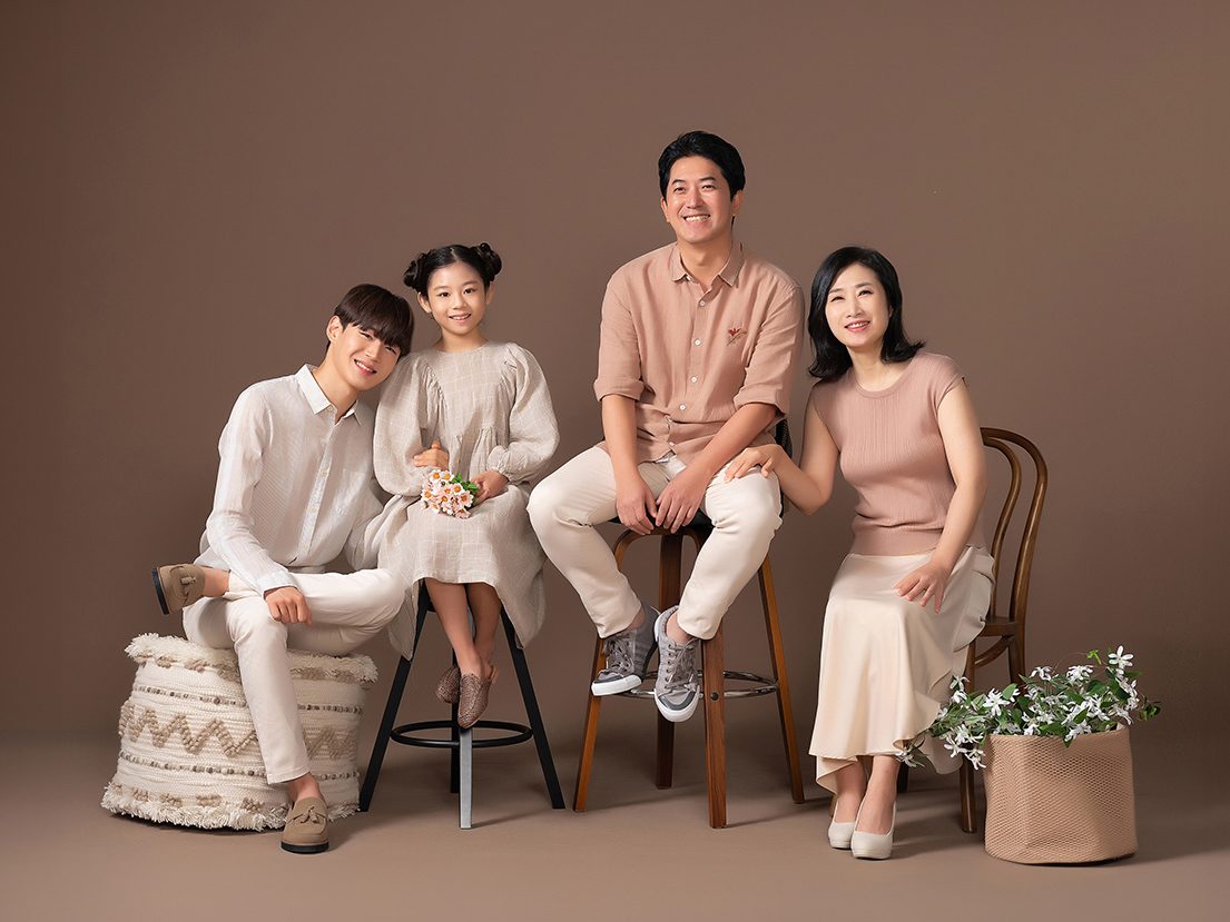창원 캐주얼 가족사진 — 갈색 배경 앞 네 식구. 부모와 아들, 어린 딸. 화이트와 연분홍이 배경과 잘 어울립니다