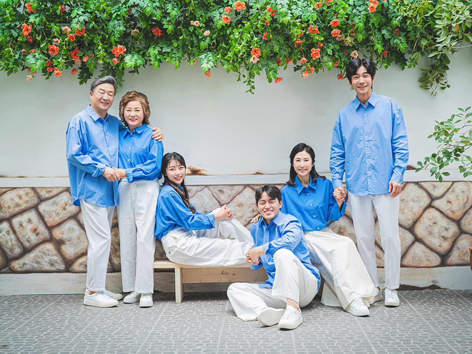 창원 캐주얼 가족사진 — 꽃 덩굴이 있는 야외 돌담 앞. 파란 셔츠와 흰 하의가 초록 배경에서 살아납니다