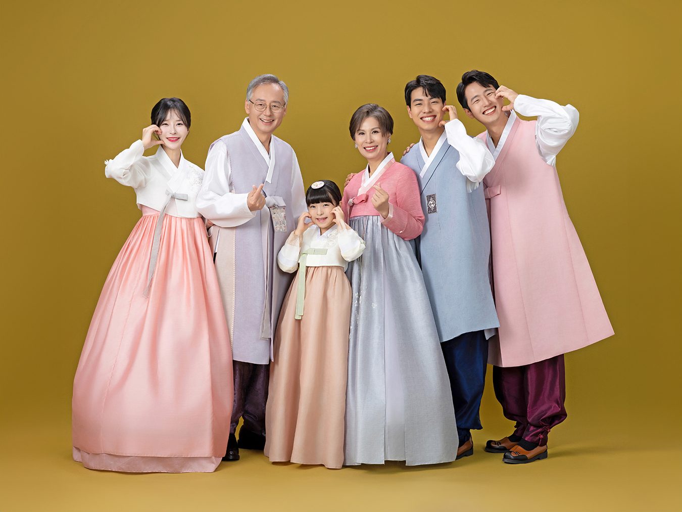 의령 한복 가족사진 — 노란 배경 앞 여섯 사람. 분홍과 연보라 저고리를 입고 두 팔을 들어 손을 흔듭니다