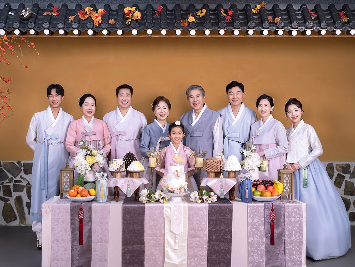 의령 한복 가족사진 — 한옥 벽 앞에 긴 상을 차리고 아홉 사람이 섰습니다. 상에는 과일과 떡, 꽃과 감이 올라가 있습니다
