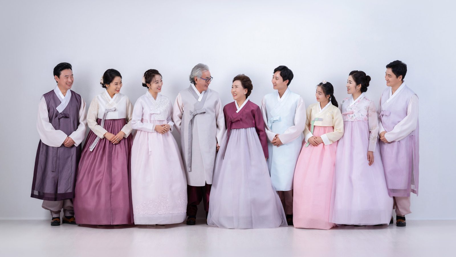 의령 한복 가족사진 — 연보라와 자주 한복을 입은 아홉 사람이 흰 배경 앞에 나란히 섰습니다