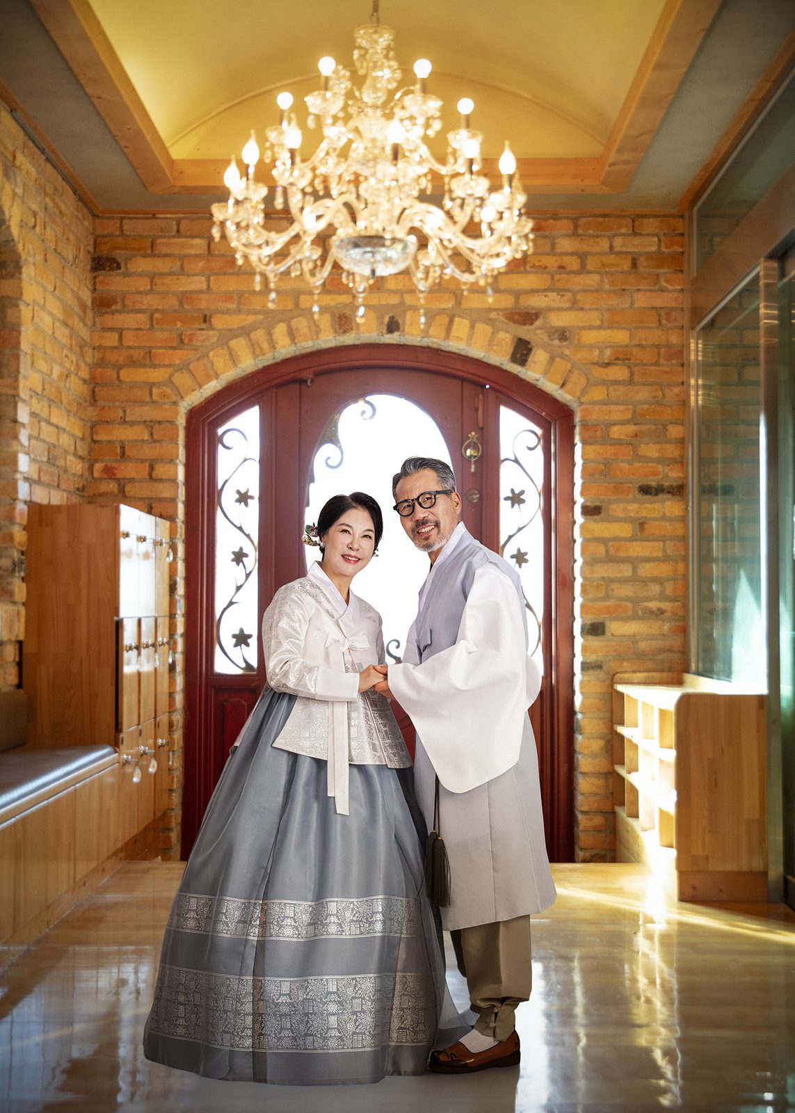 창원 한복 부부사진 — 벽돌 벽과 붉은 문, 샹들리에 아래에 선 두 사람. 아내는 회색 치마, 남편은 베이지 두루마기입니다