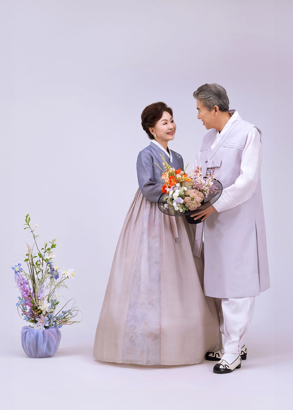 창원 한복 부부사진 — 연보라 배경 앞에서 꽃바구니를 든 아내와 남편. 두 사람이 서로를 보고 웃습니다