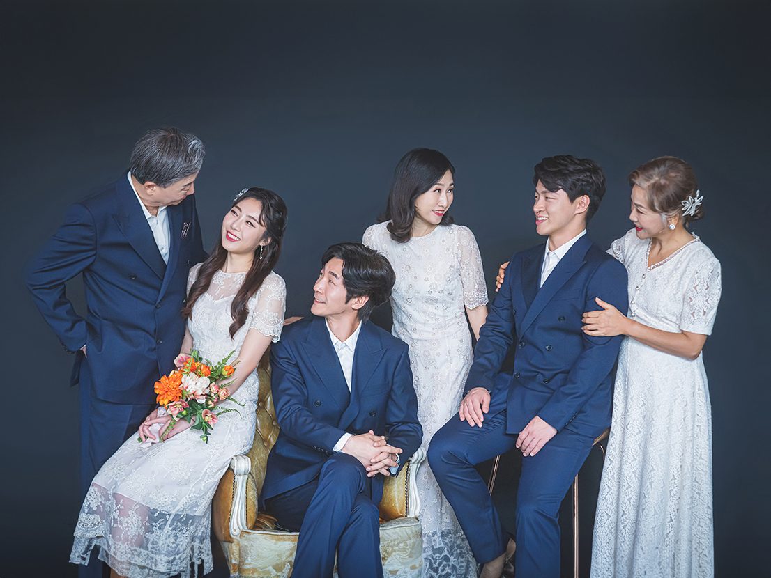 창원 세미웨딩 가족사진 — 어두운 회색 배경 앞에서 서로를 바라보는 여섯 사람. 배경이 어두우면 흰 드레스가 더 밝게 보입니다