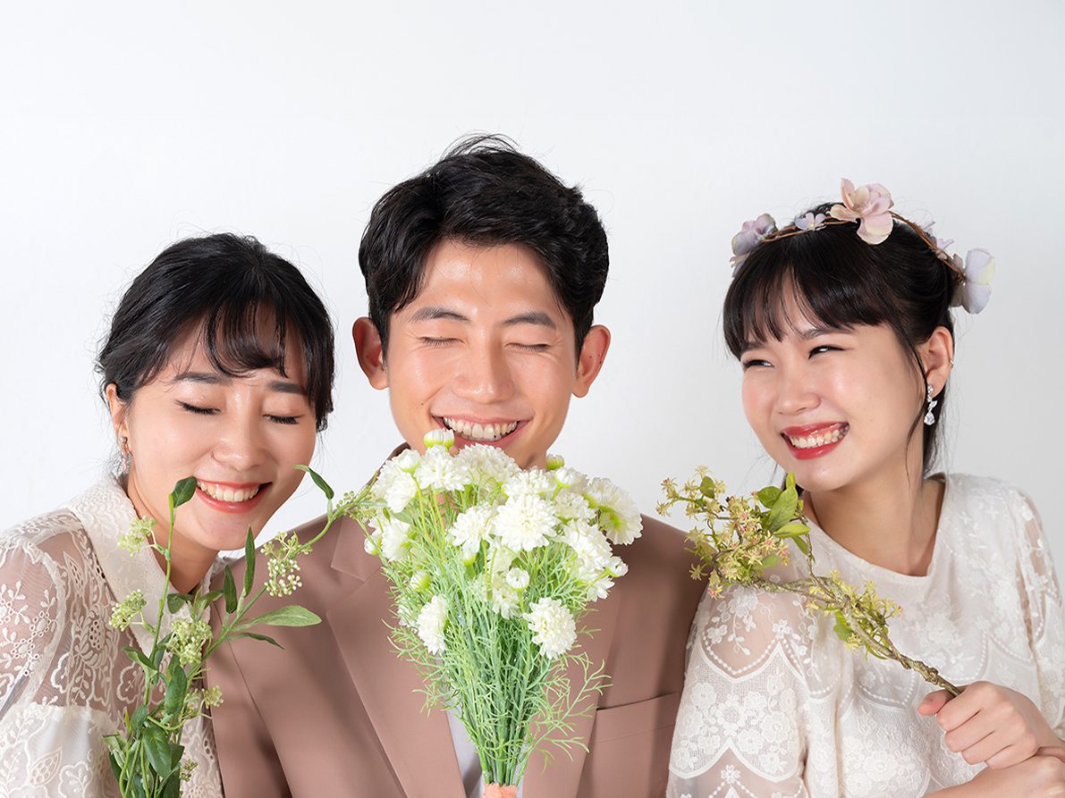 창원 세미웨딩 가족사진 — 갈색 수트를 입은 청년과 흰 레이스 원피스를 입은 두 여성. 꽃을 들고 가까이 섰습니다