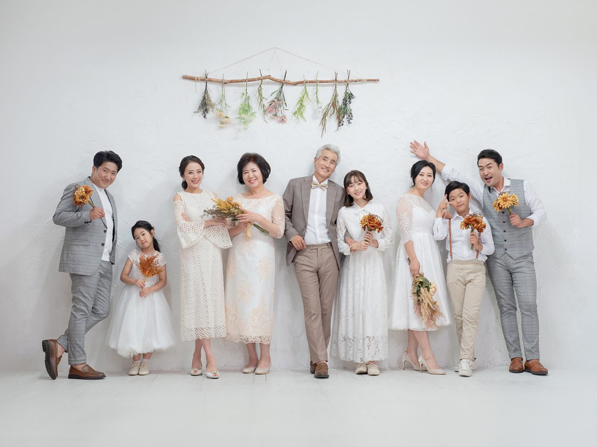 창원 세미웨딩 가족사진 — 마른 꽃을 건 흰 벽 앞에서 여덟 사람. 꽃다발이 계절감을 만듭니다