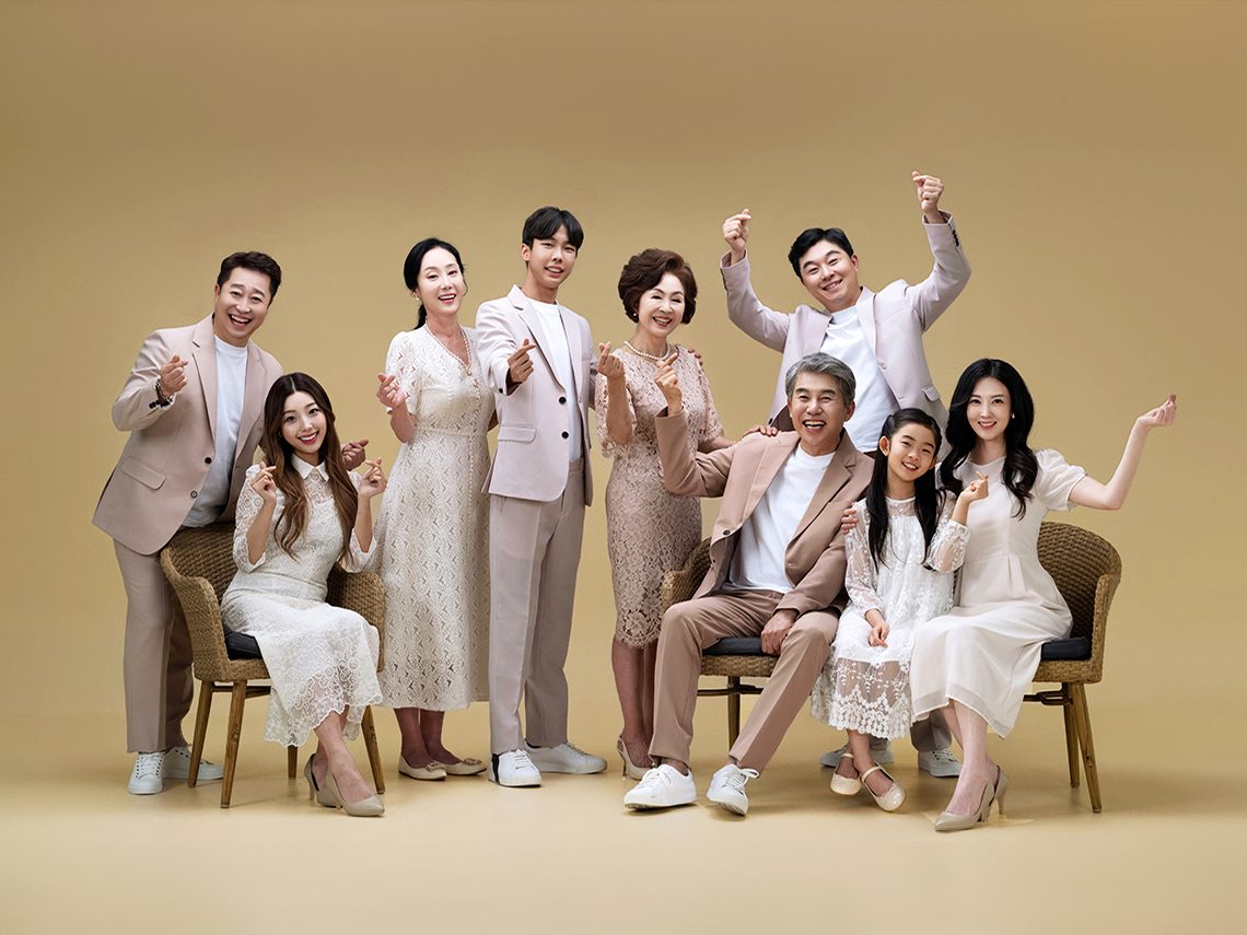 창원 세미웨딩 가족사진 — 베이지 배경 앞 아홉 사람. 손가락 하트와 뻗은 팔이 사진을 밝게 만듭니다